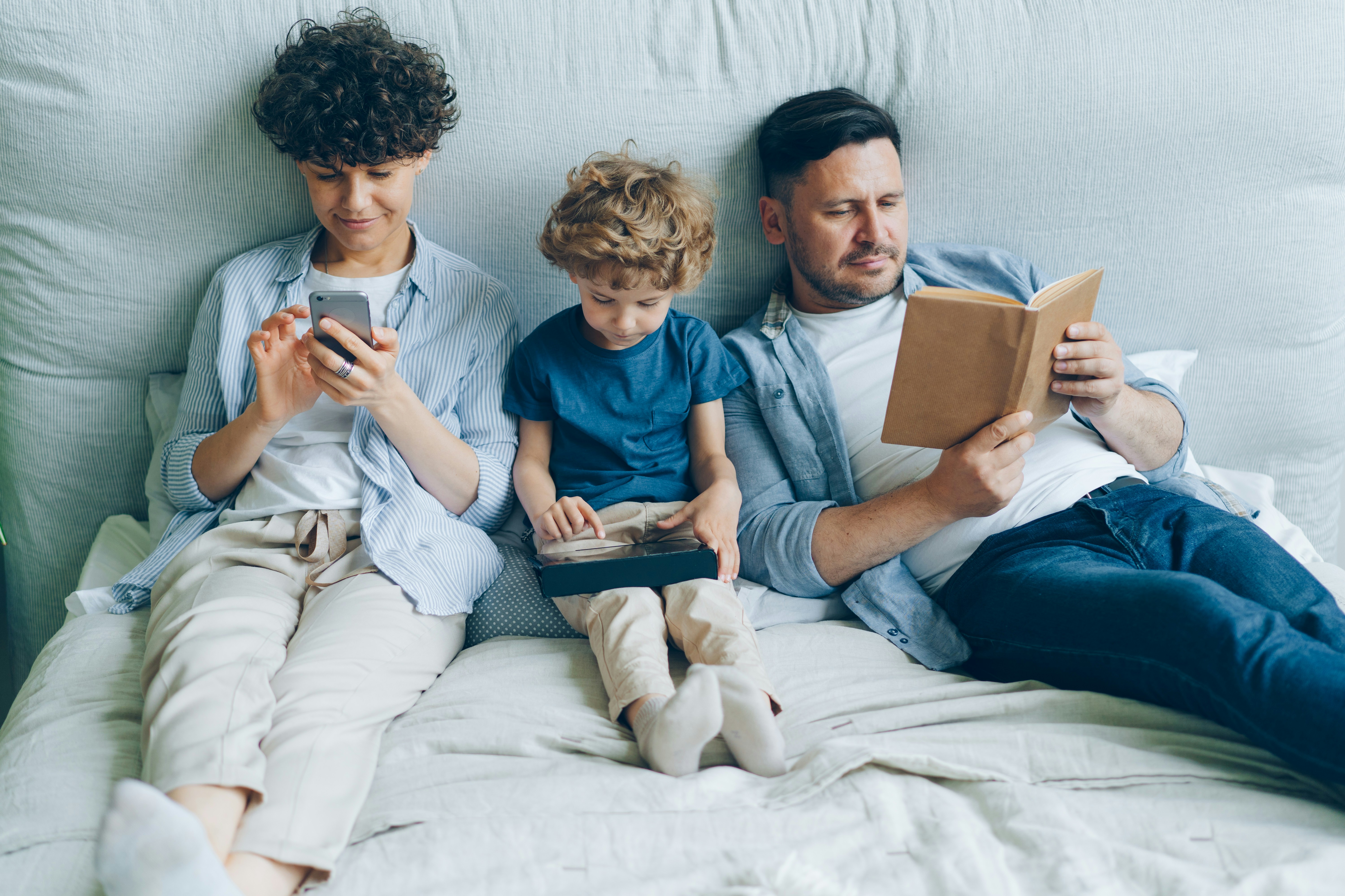 Mother using smartphone son holding tablet father reading book in bed at home